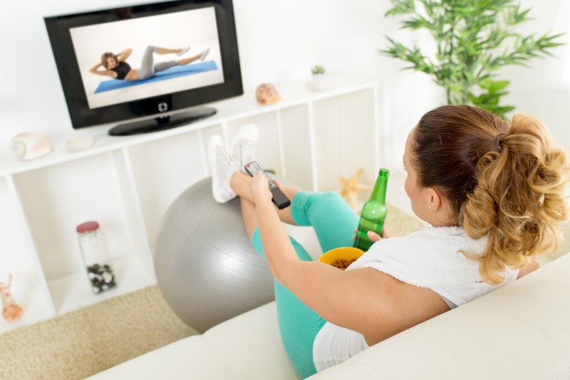 Frau sitzt entspannt auf Sofa mit Bier und Essen und schaut Home-Workout im Fernsehen